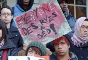 group of protesters holding a sign that reads "everyone deserves a bed"